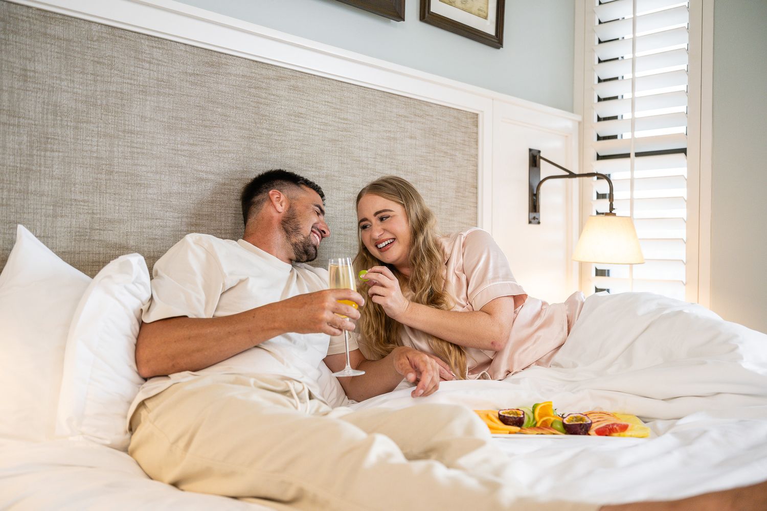Couple enjoying breakfast in bed, toasting with champagne glasses in a bright, elegant hotel room.