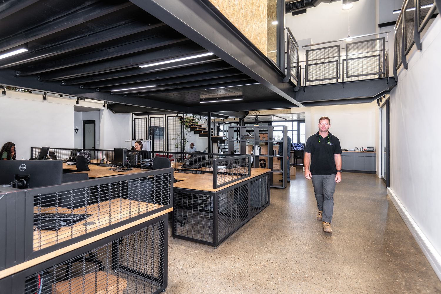 Modern open-plan office with industrial black metal workstations, mezzanine level, and polished concrete floors.