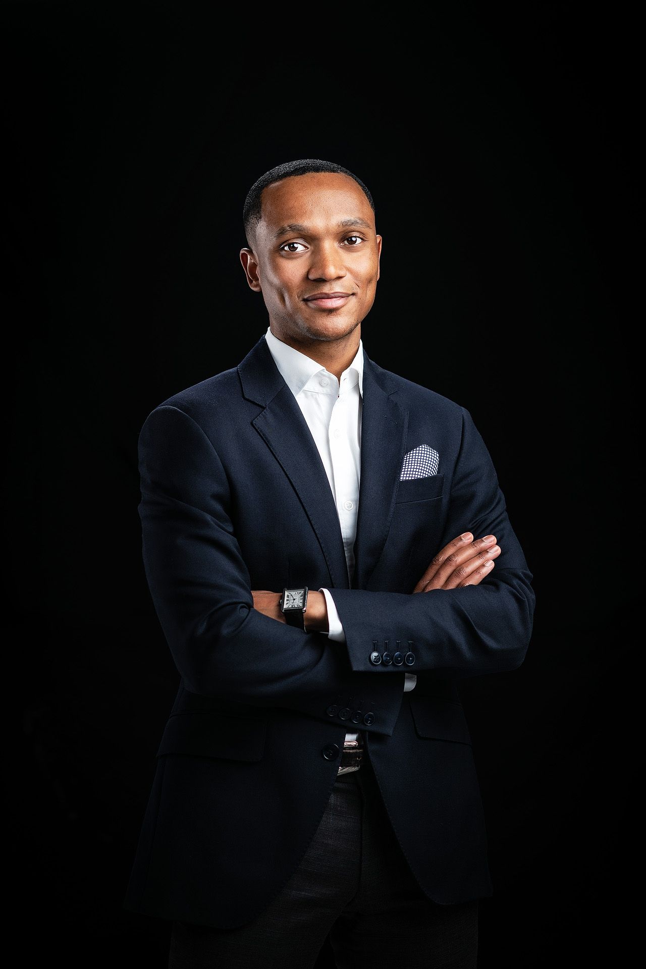 Professional Black man in navy suit, arms crossed, smiling confidently against black background.