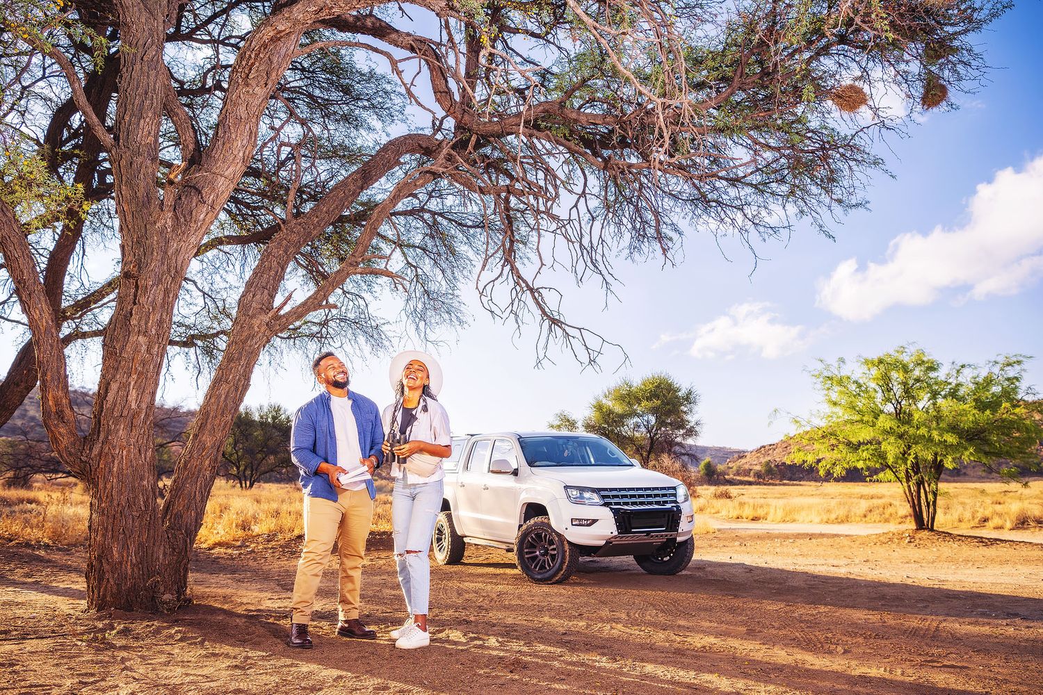 Two people stand under an acacia tree beside a white SUV in an African savanna landscape on a sunny day.