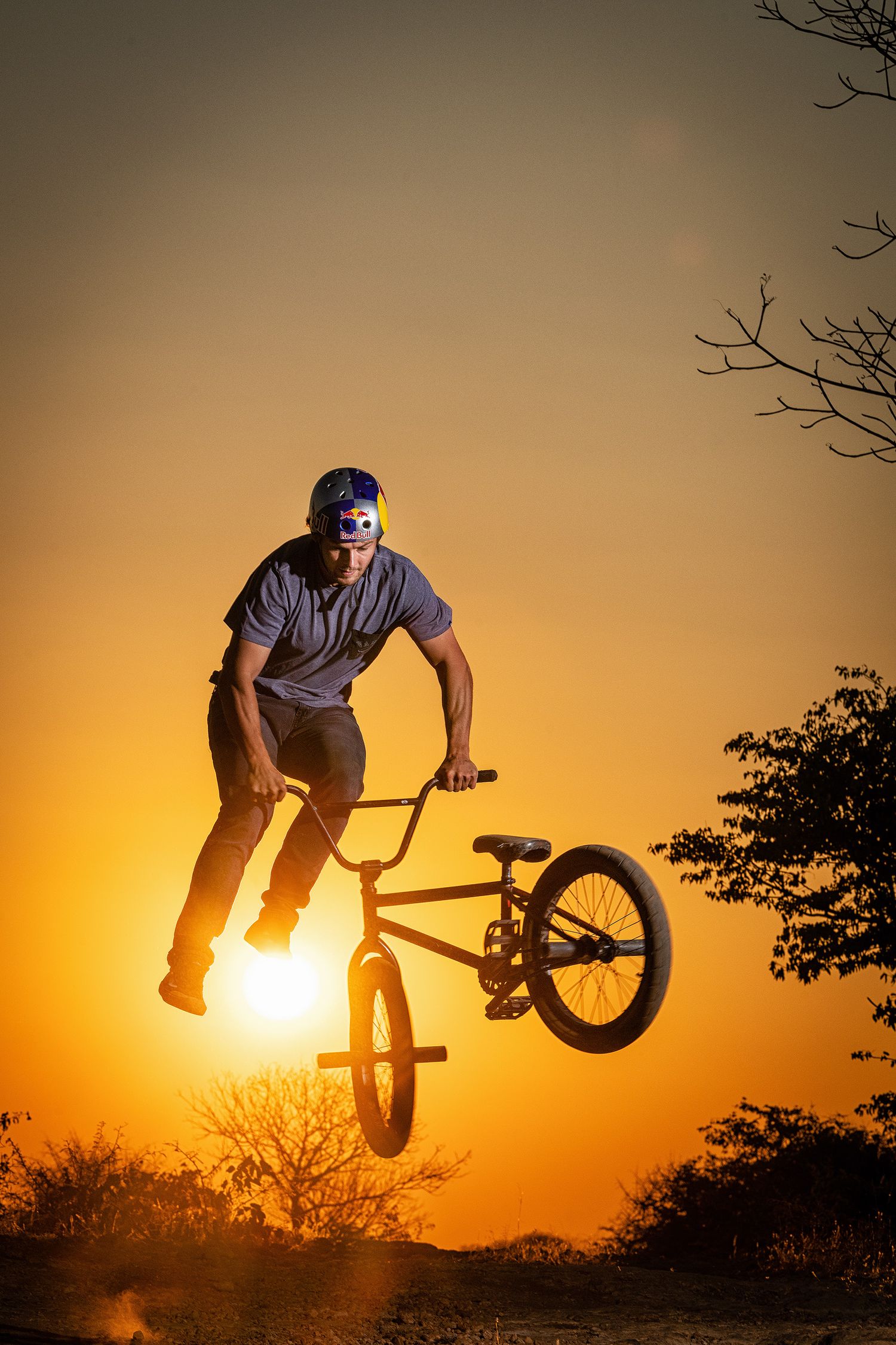 BMX rider performs a dramatic aerial trick silhouetted against a vibrant orange sunset sky with bare trees in background.