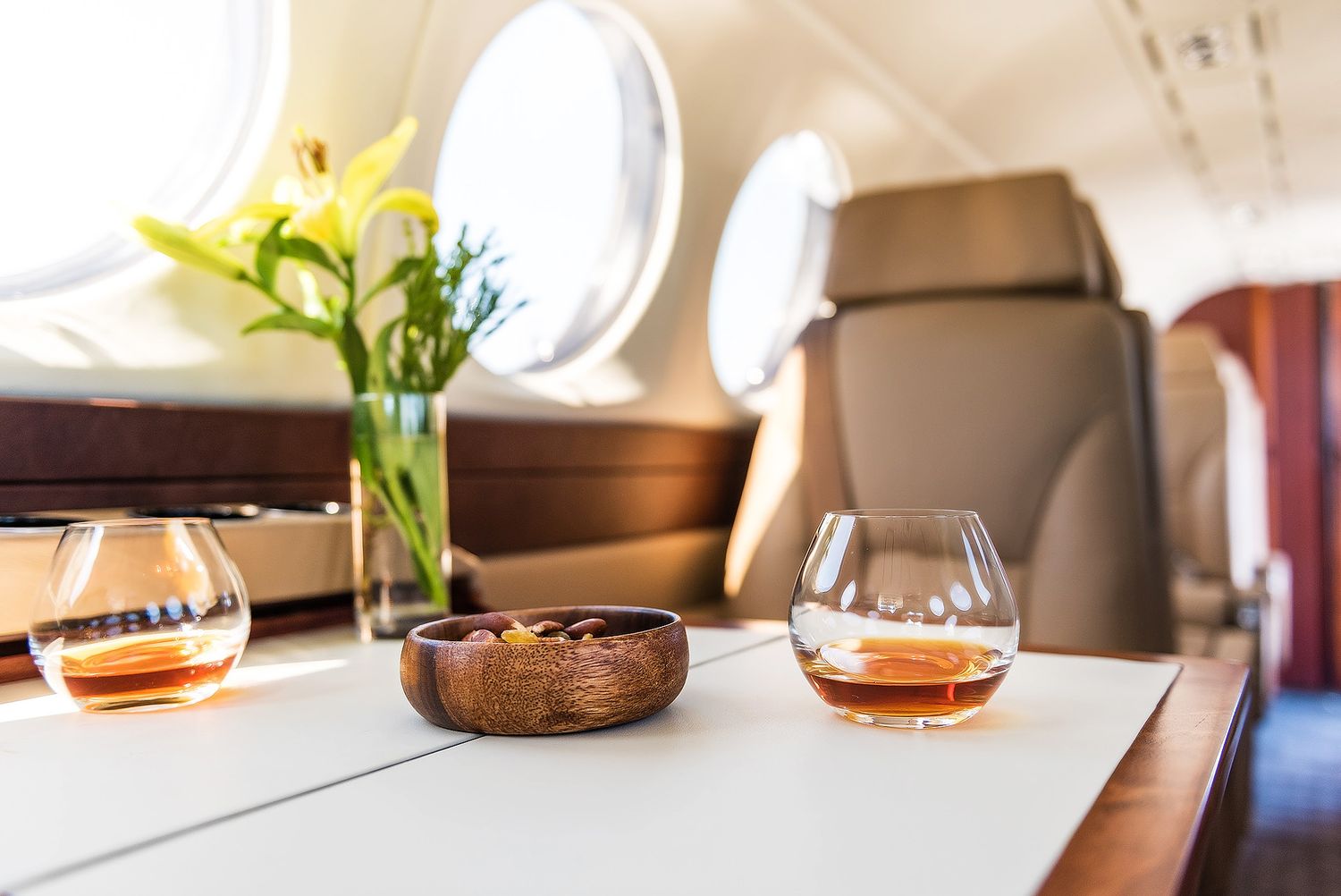 Luxury private jet interior with whiskey glasses, wooden bowl, and yellow lily flowers on a white table near oval windows.