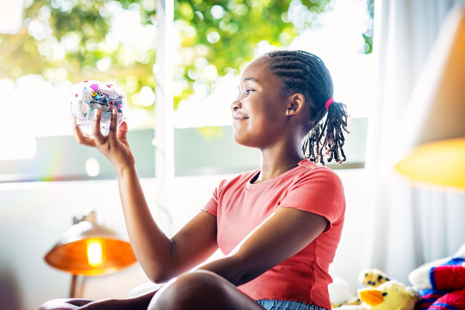 Young Black girl smiling while holding a colorful glitter jar near a bright window in a cozy room.