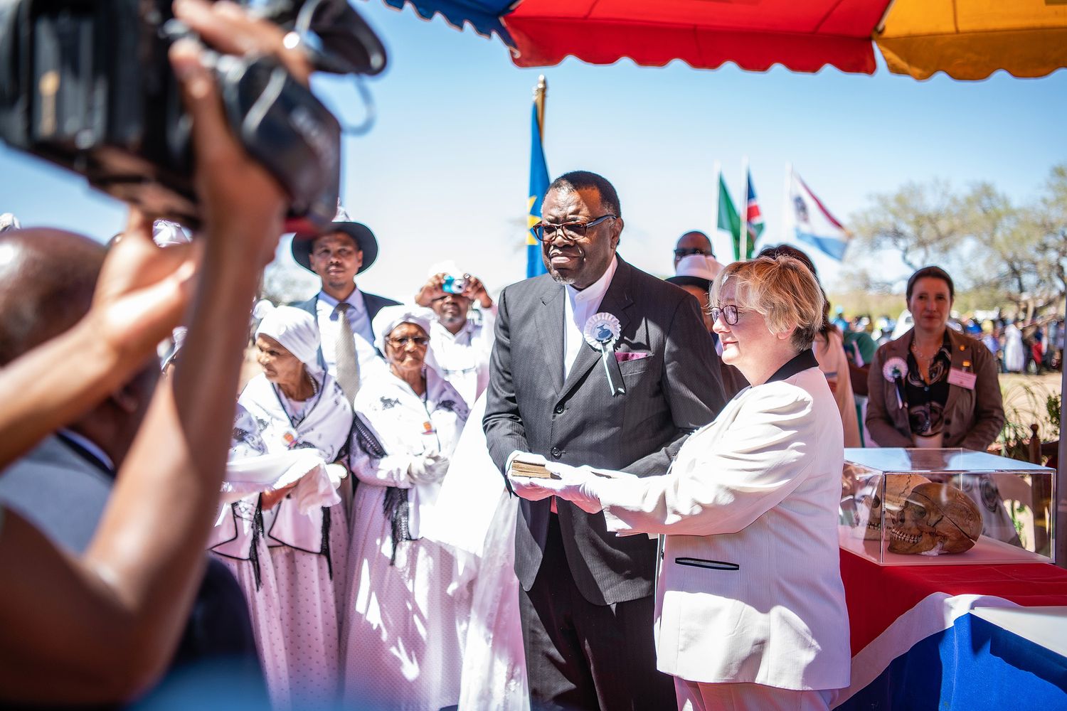 Namibian official presenting a certificate at an outdoor community ceremony with colorful flags and crowd.