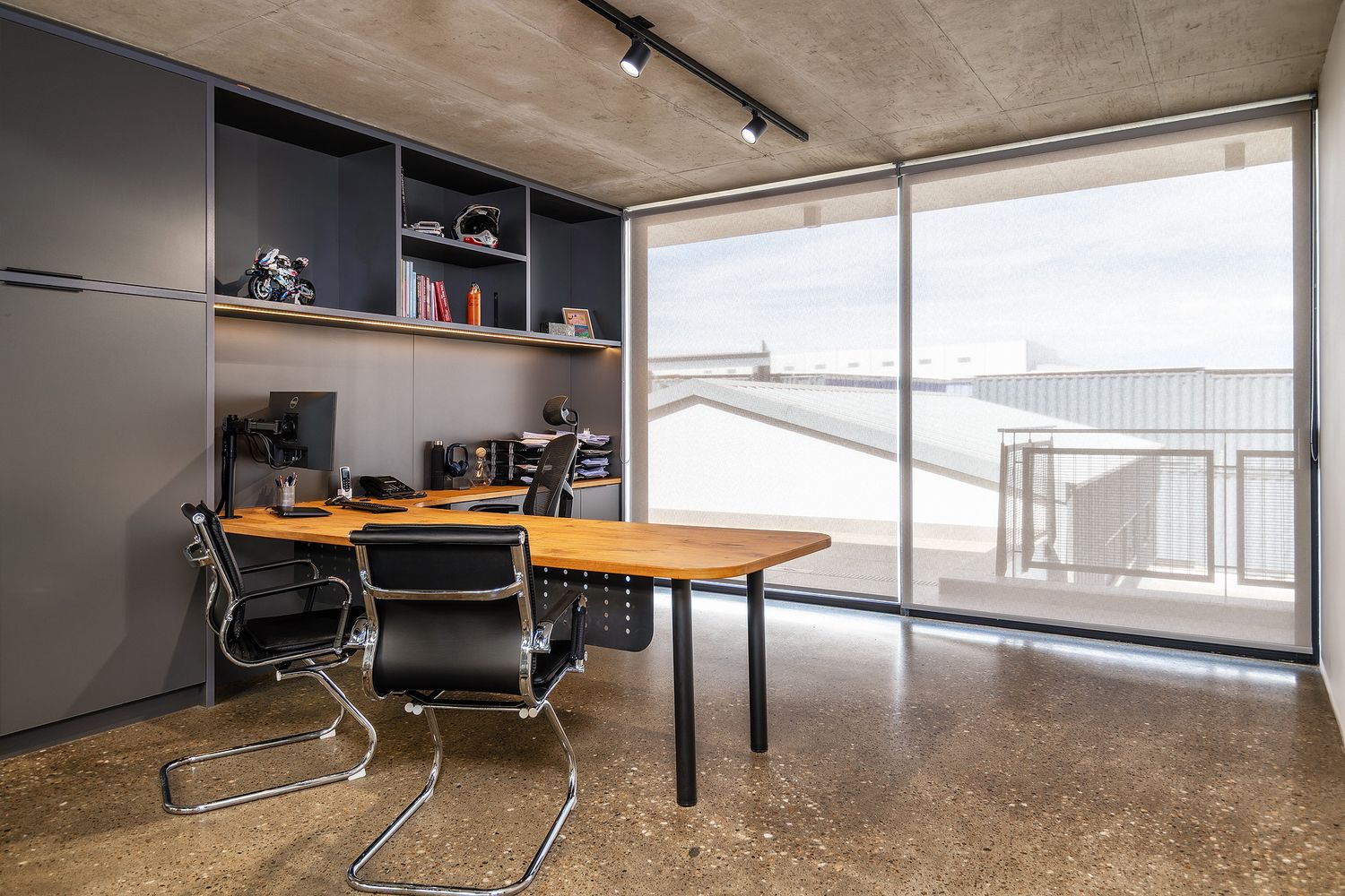 Modern office with wooden desk, ergonomic chairs, built-in shelving, and large window overlooking a balcony.