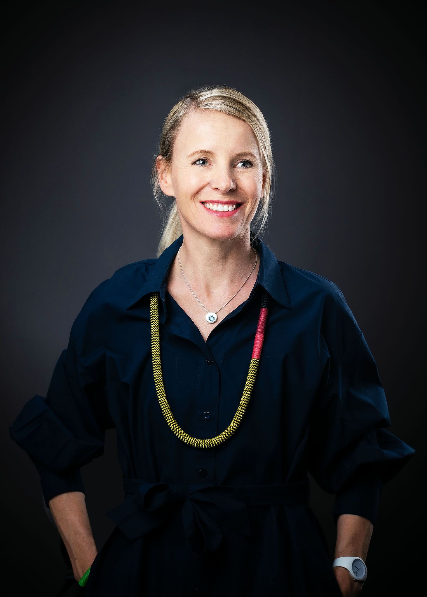 Smiling blonde woman in navy shirt with colorful necklace poses confidently against dark background.