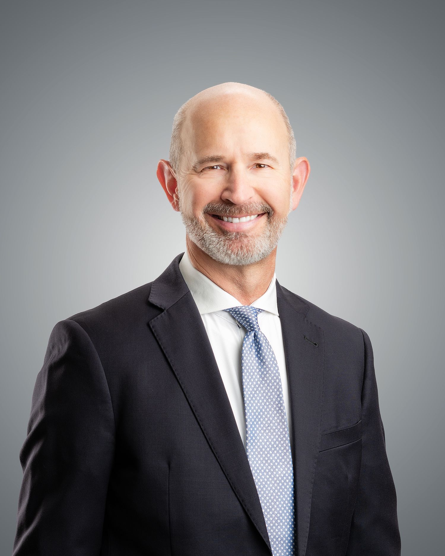 Professional headshot of a smiling bald man with a gray beard wearing a dark suit and light blue tie.