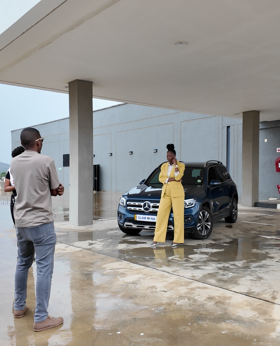 Woman in yellow jumpsuit poses beside a Mercedes-Benz SUV while a photographer captures the shot outdoors.