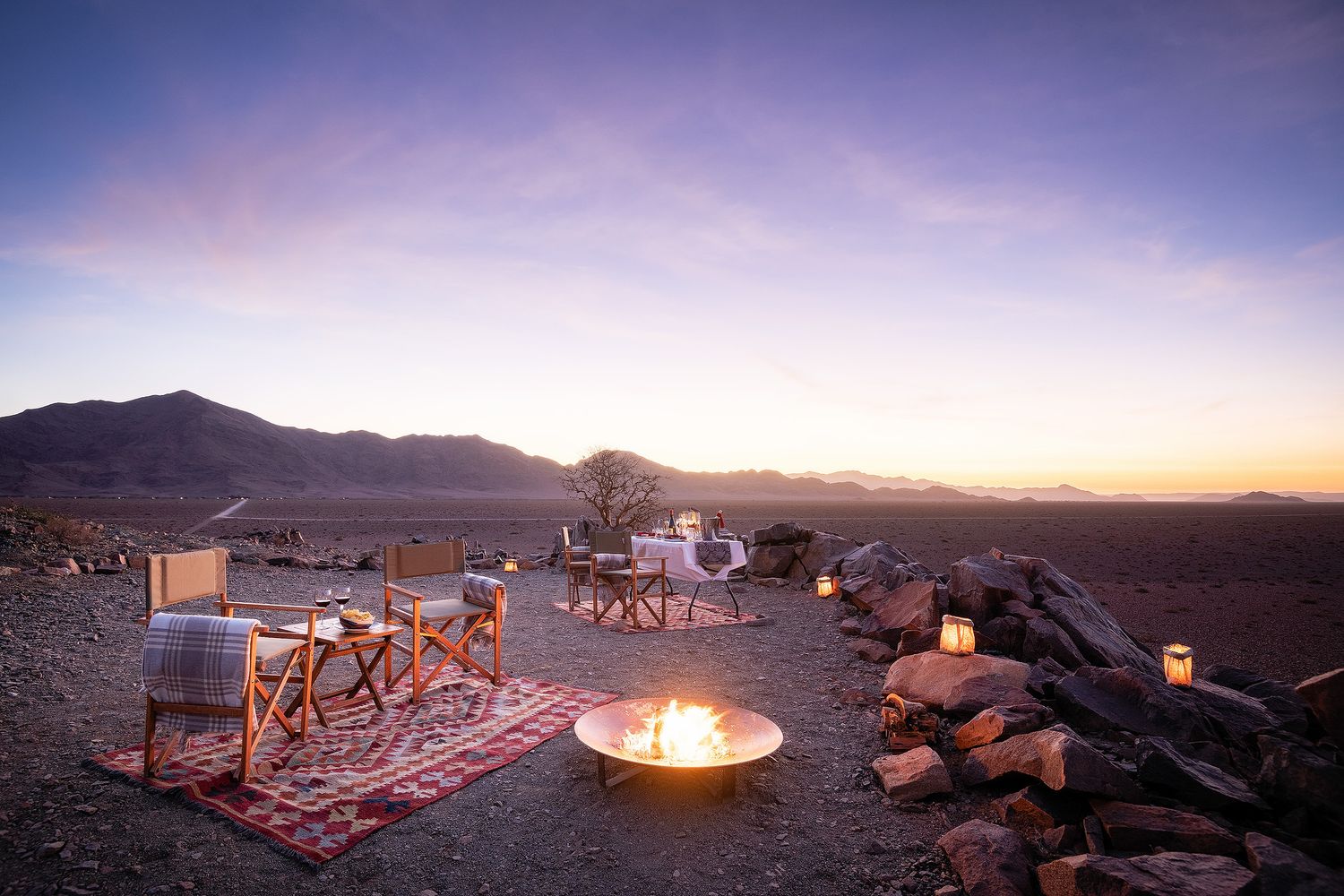 Desert campfire setup at dusk with chairs, rugs, and lanterns against a mountain backdrop under a colorful twilight sky.
