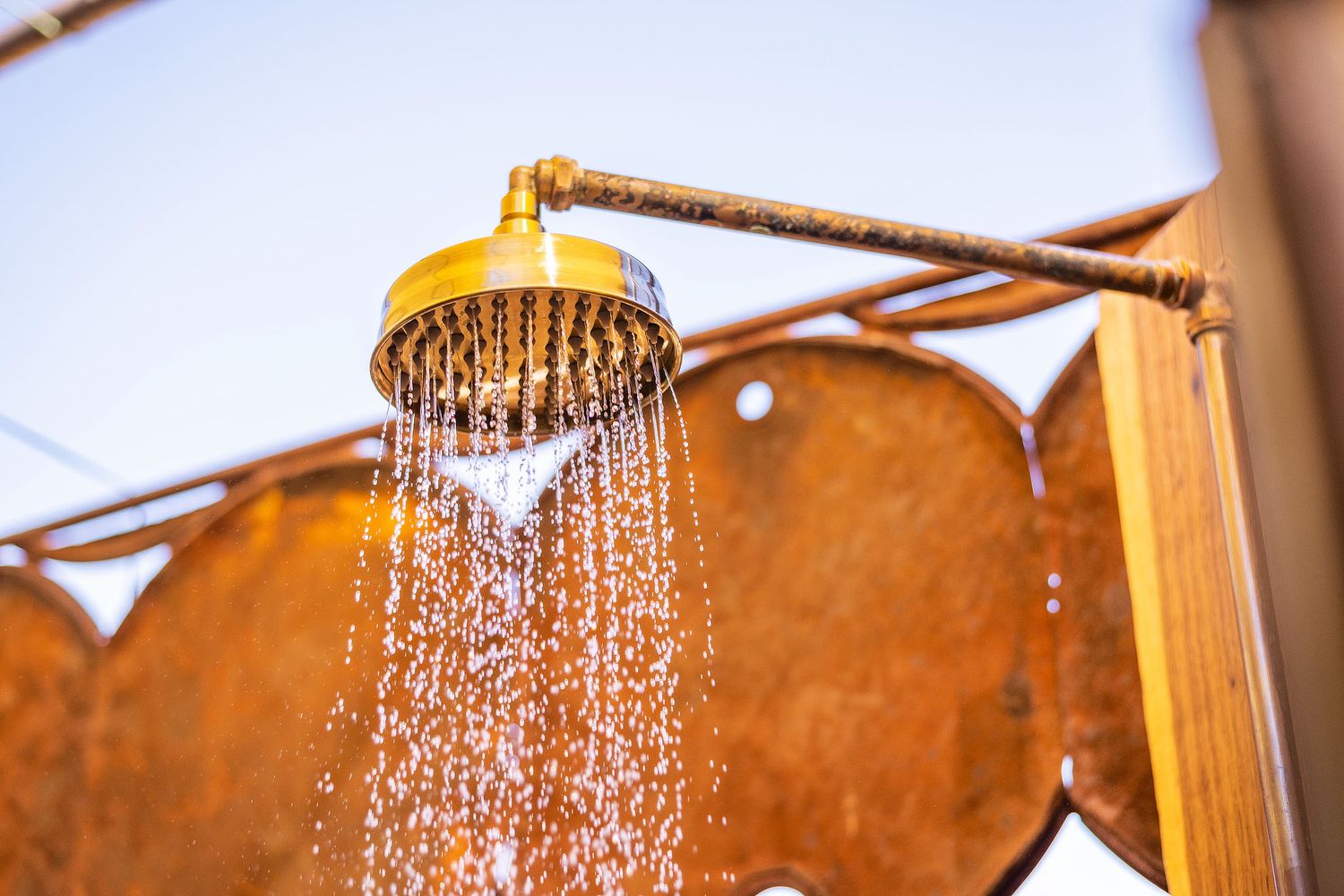 Golden outdoor shower head spraying water against a clear blue sky with rustic wooden backdrop.