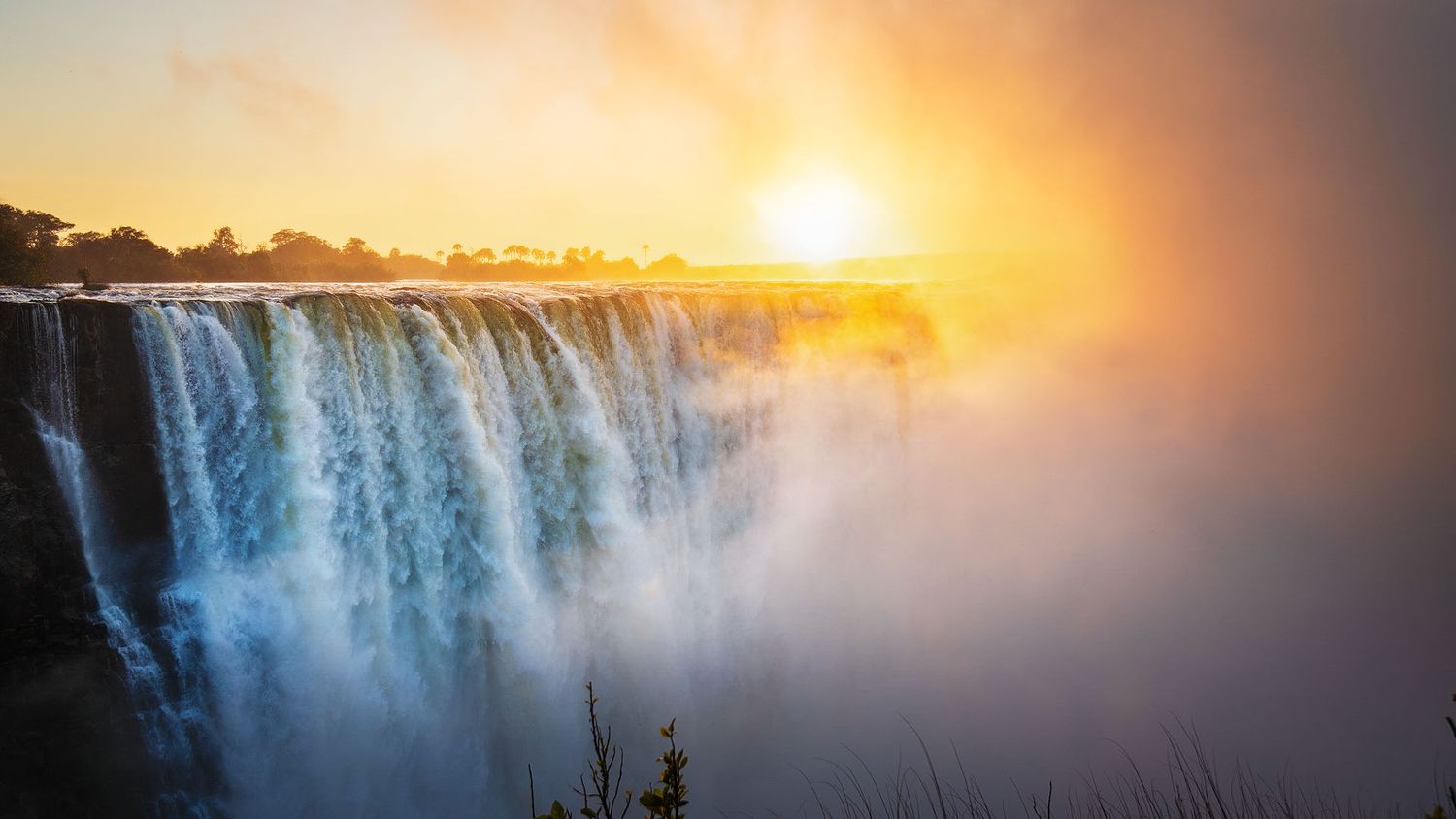 Stunning sunset over Victoria Falls with golden light illuminating cascading waterfalls and rising mist.
