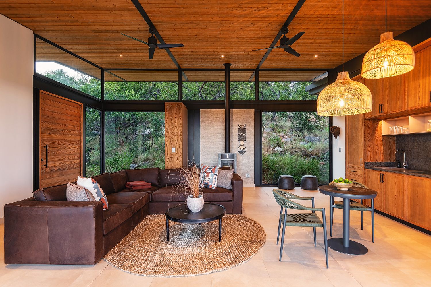 Modern safari lodge interior with leather sectional sofa, rattan pendant lights, and floor-to-ceiling windows overlooking lush greenery.
