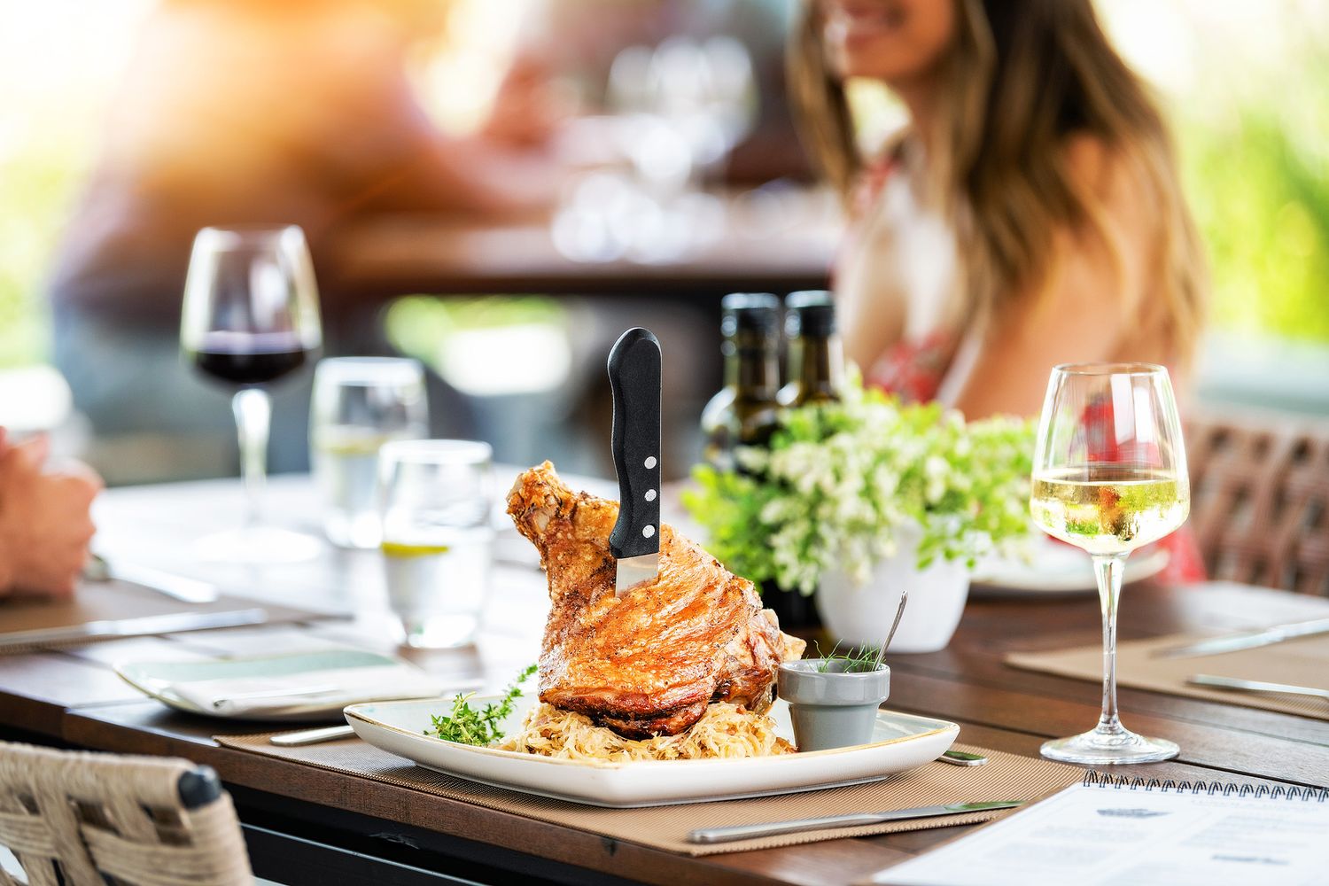 Elegant restaurant table with roasted chicken dish, wine glasses, and blurred diners in a bright outdoor setting.