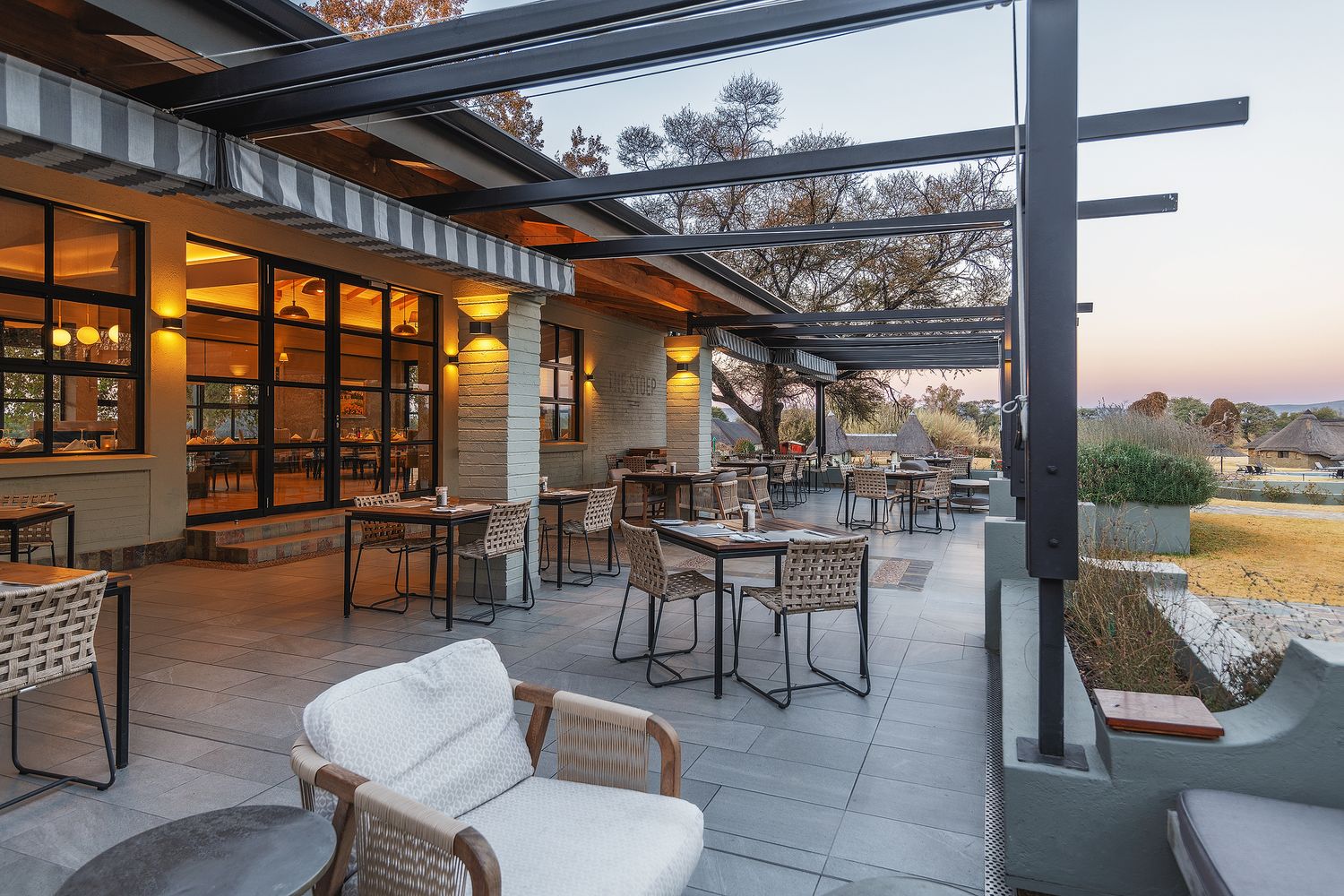 Elegant outdoor patio with wicker chairs, dining tables, and pergola at dusk overlooking a scenic landscape.
