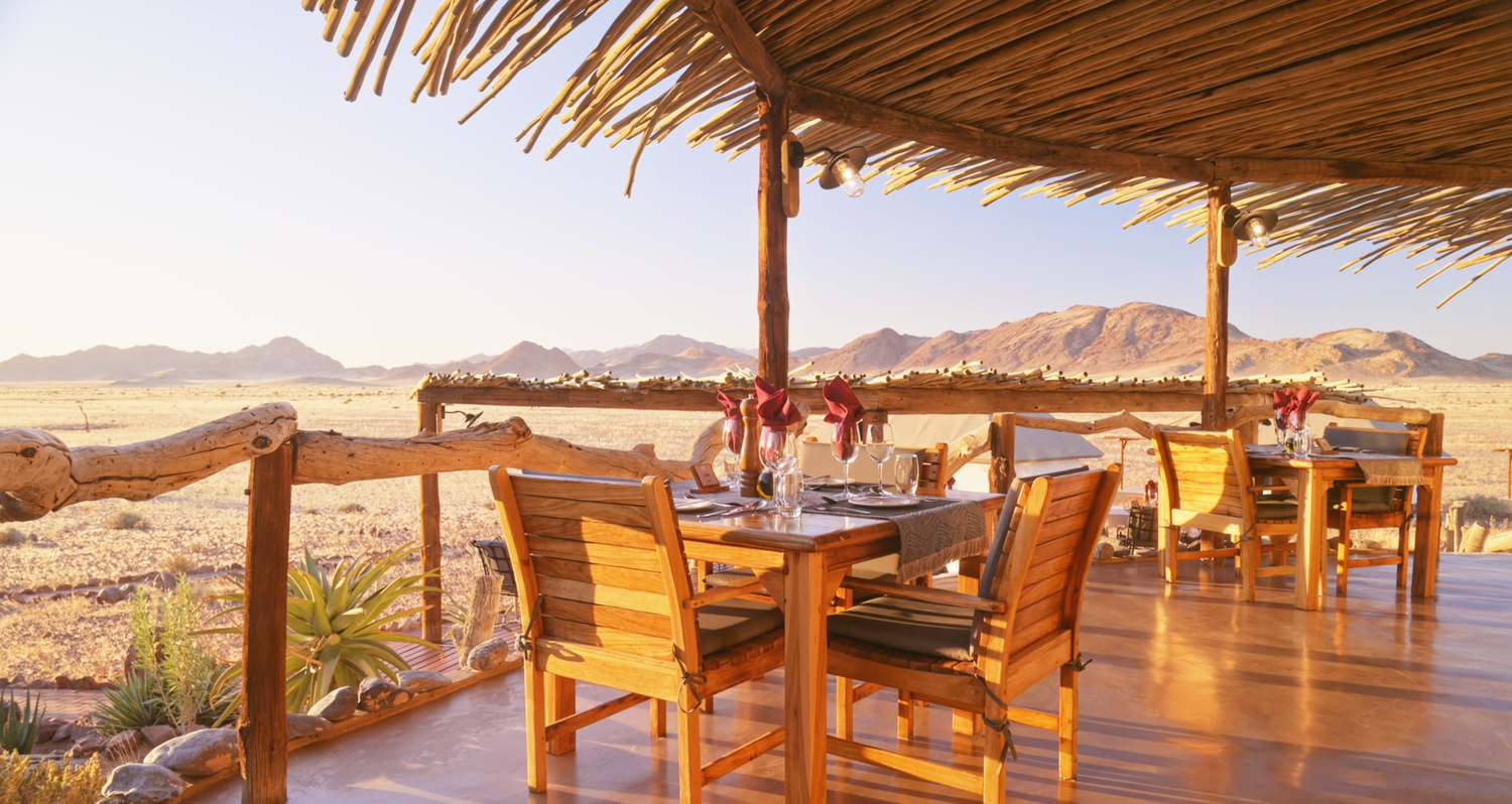 Outdoor desert lodge dining area with wooden furniture, thatched roof, and stunning mountain landscape views.