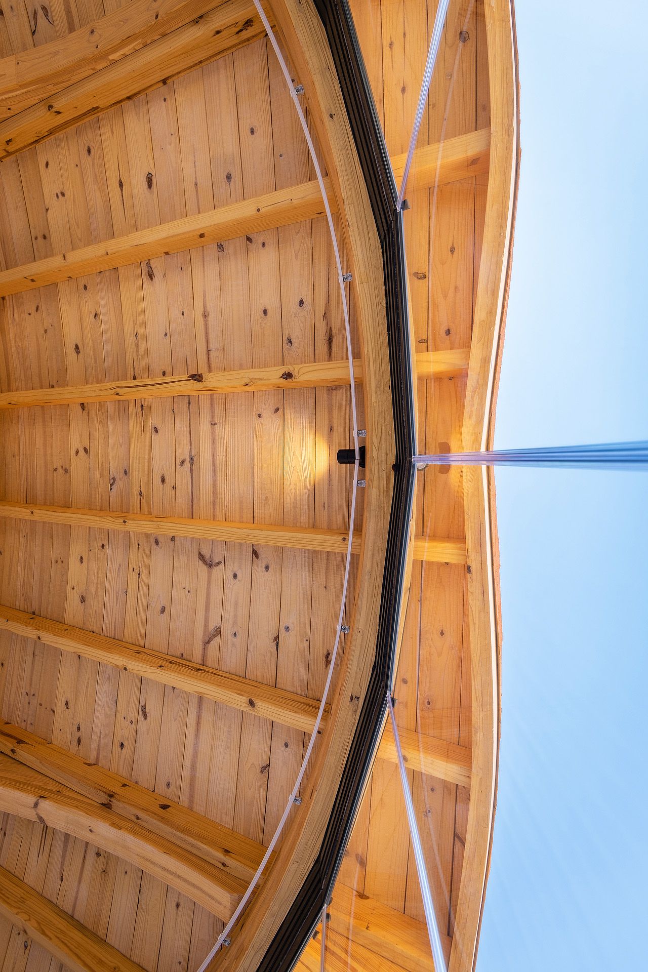 Looking up at a curved wooden ceiling with radial beams and a central metal pole against blue sky.