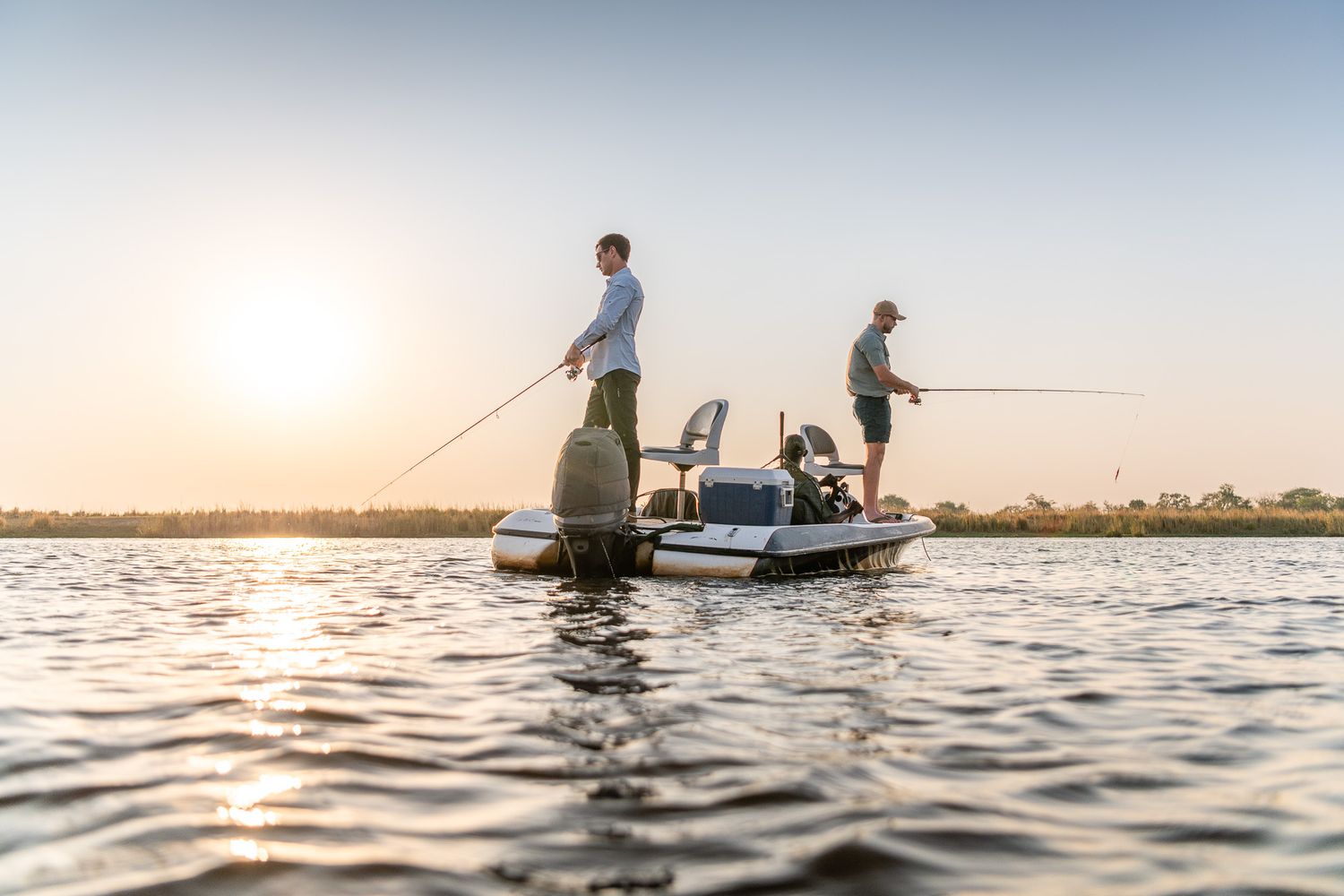 Two men fishing from a small boat on calm water at sunset with fishing rods cast out.