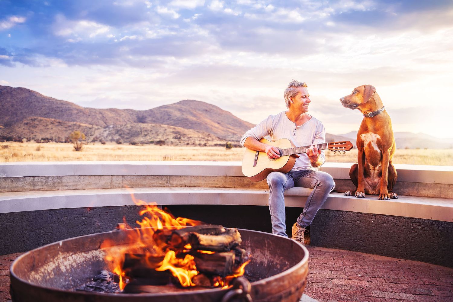 Man playing guitar by a fire pit with a dog, mountains and sunset sky in the background.
