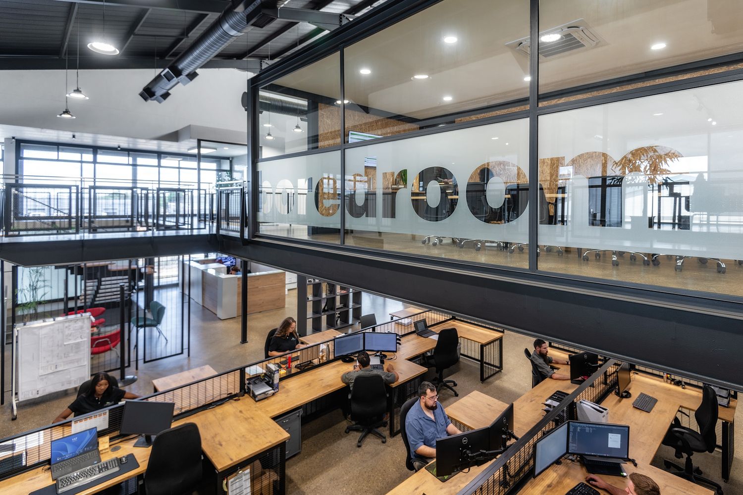 Modern open-plan office with employees working at wooden desks, large glass windows, and a bold branded logo wall.