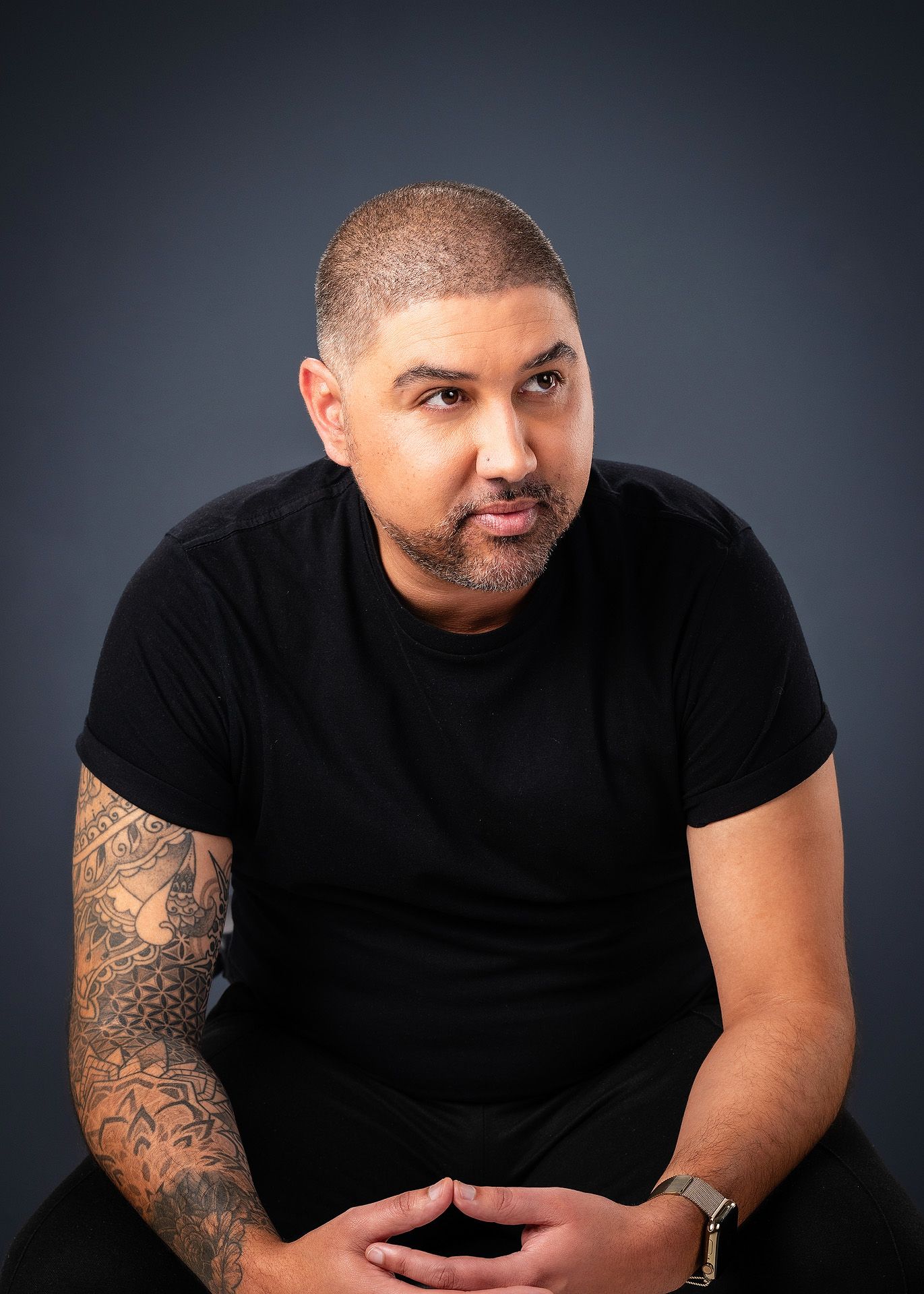 Tattooed man in black t-shirt with short hair and beard sits confidently against gray studio background.