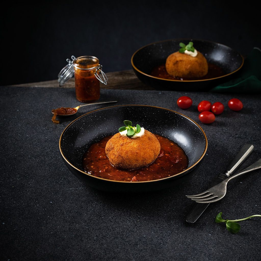 Gourmet arancini rice ball in rich tomato sauce served in elegant black bowl with cherry tomatoes.