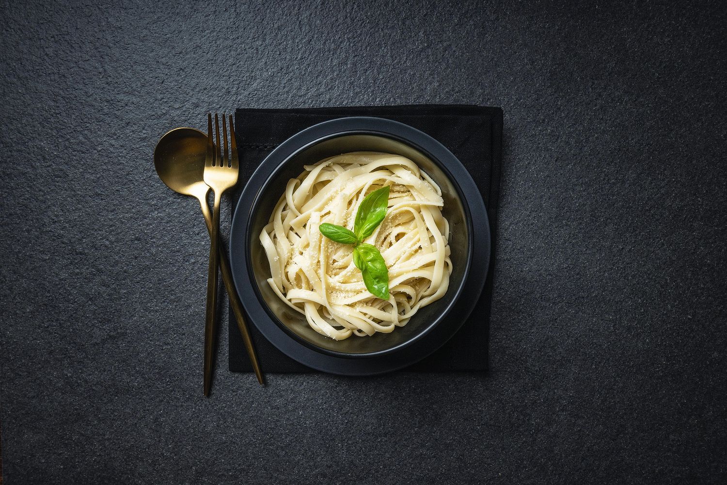 A bowl of creamy fettuccine pasta garnished with fresh basil, served on a dark plate with chopsticks and a gold spoon.