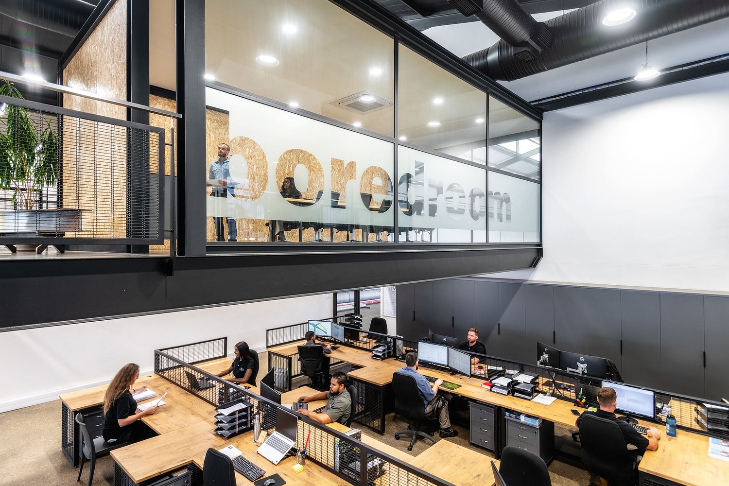 Modern open-plan office workspace with employees at desks, industrial design, large windows, and a branded logo on glass wall.
