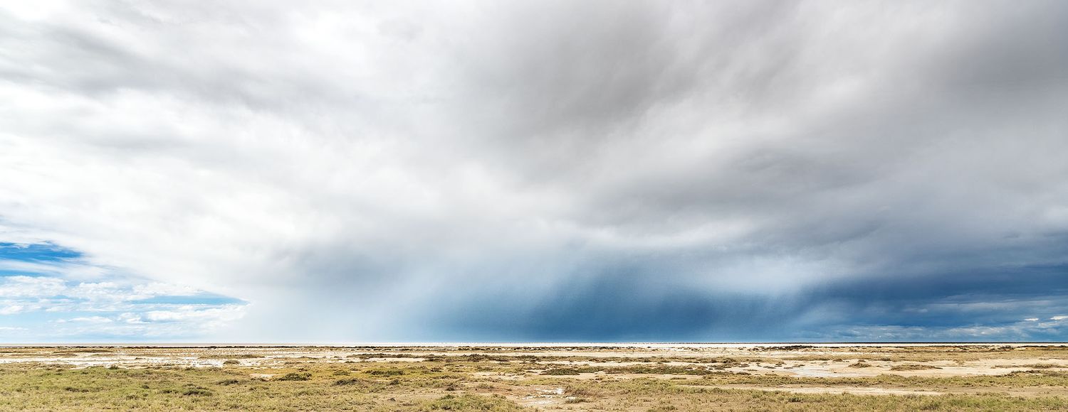 Dramatic storm clouds loom over a vast flat desert landscape under a wide open sky.