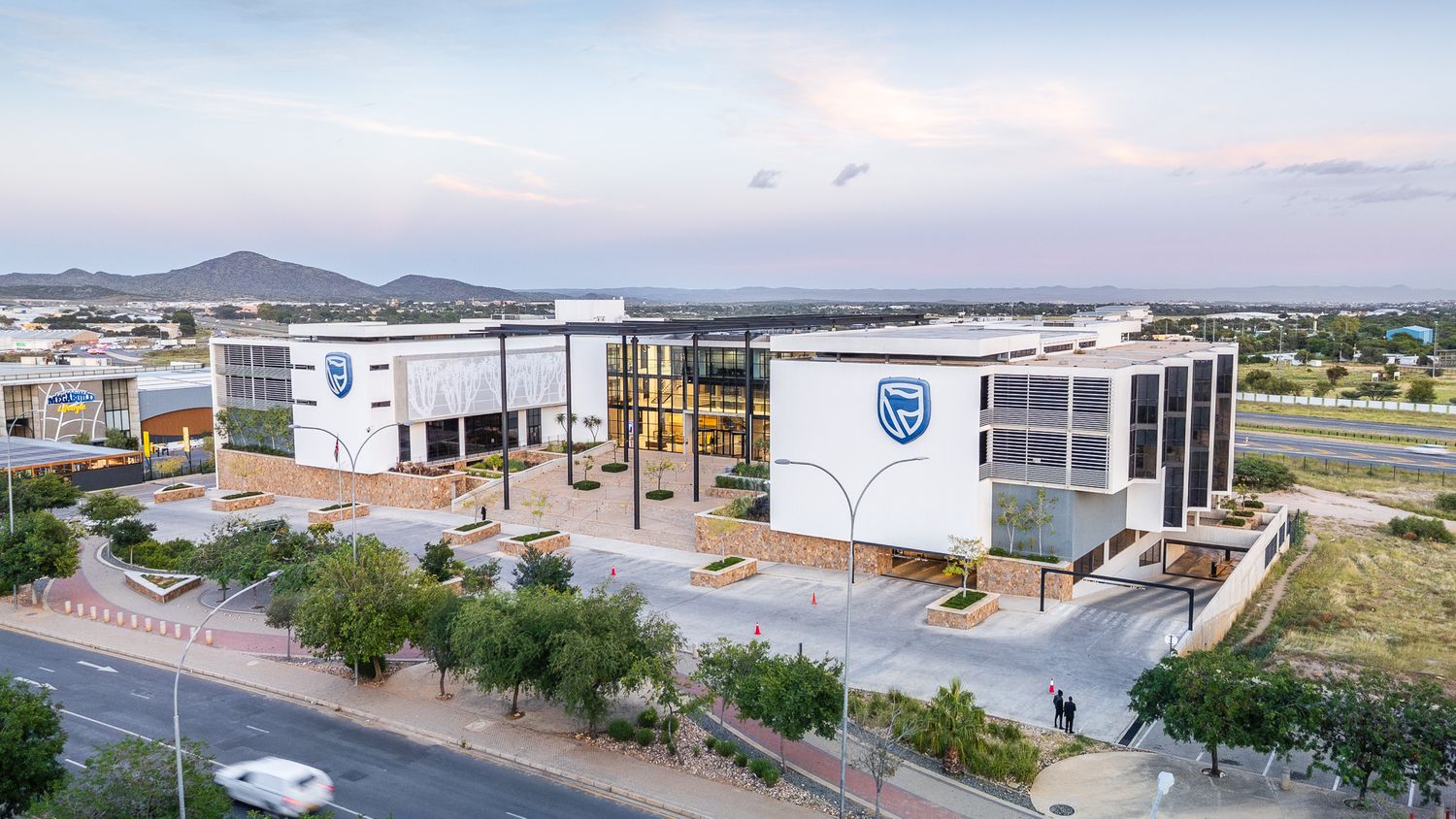 Aerial view of a modern corporate building complex with blue and white facade, surrounded by trees and mountains at dusk.