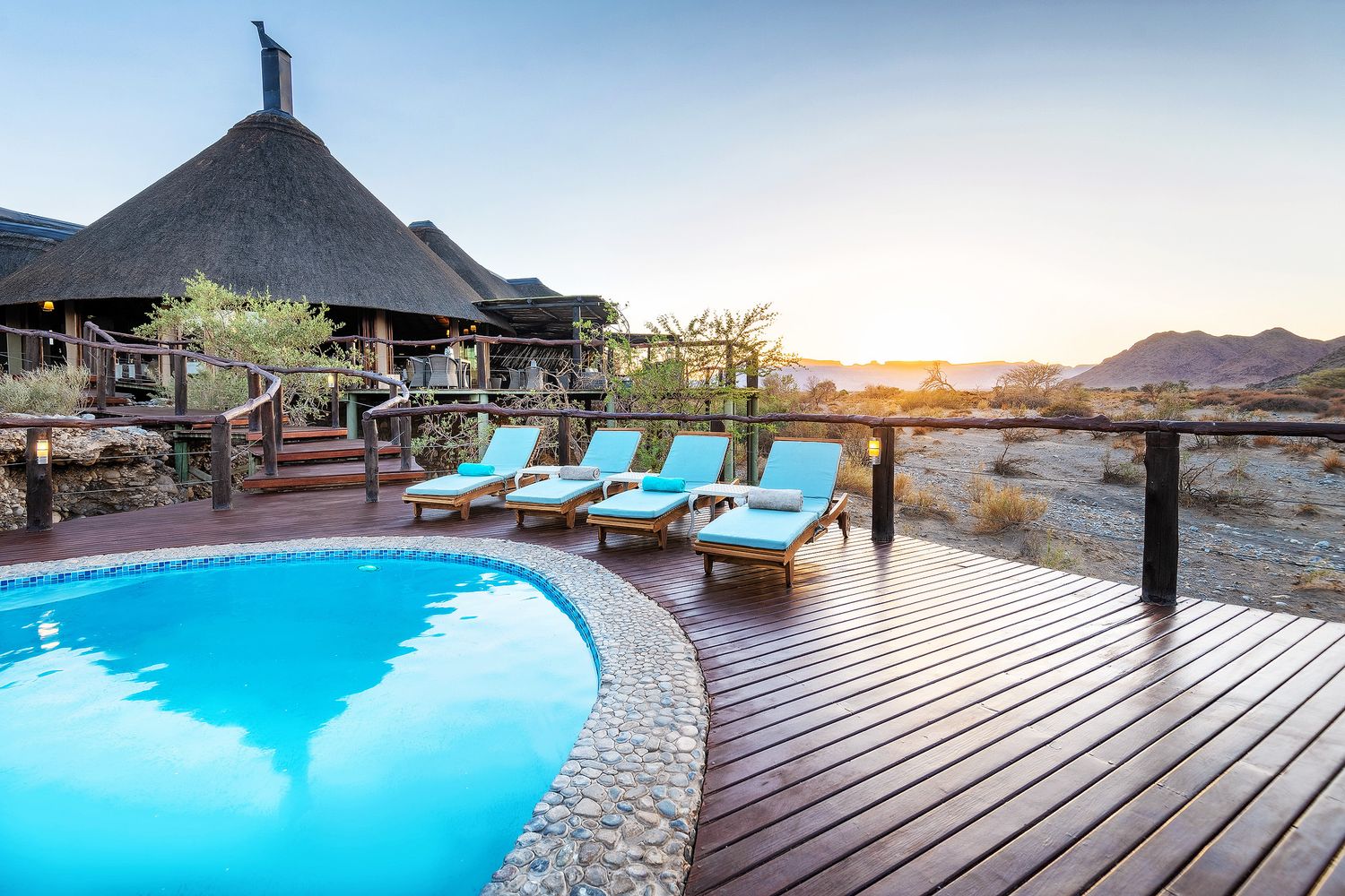 Luxury safari lodge pool with turquoise water and sun loungers overlooking a desert landscape at sunset.