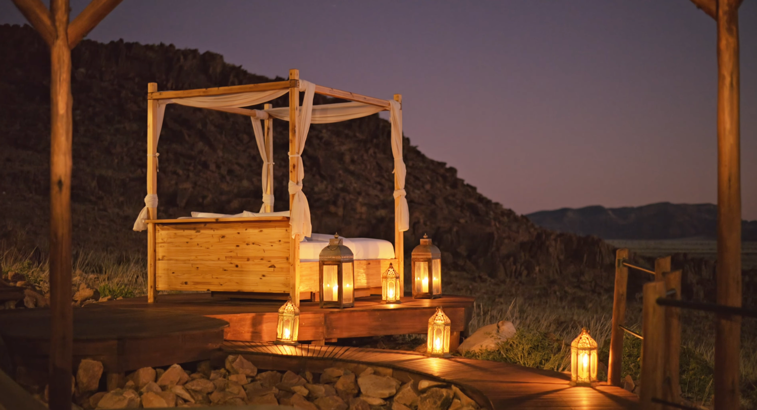 Romantic outdoor canopy bed on a wooden deck surrounded by glowing lanterns at dusk in a desert landscape.