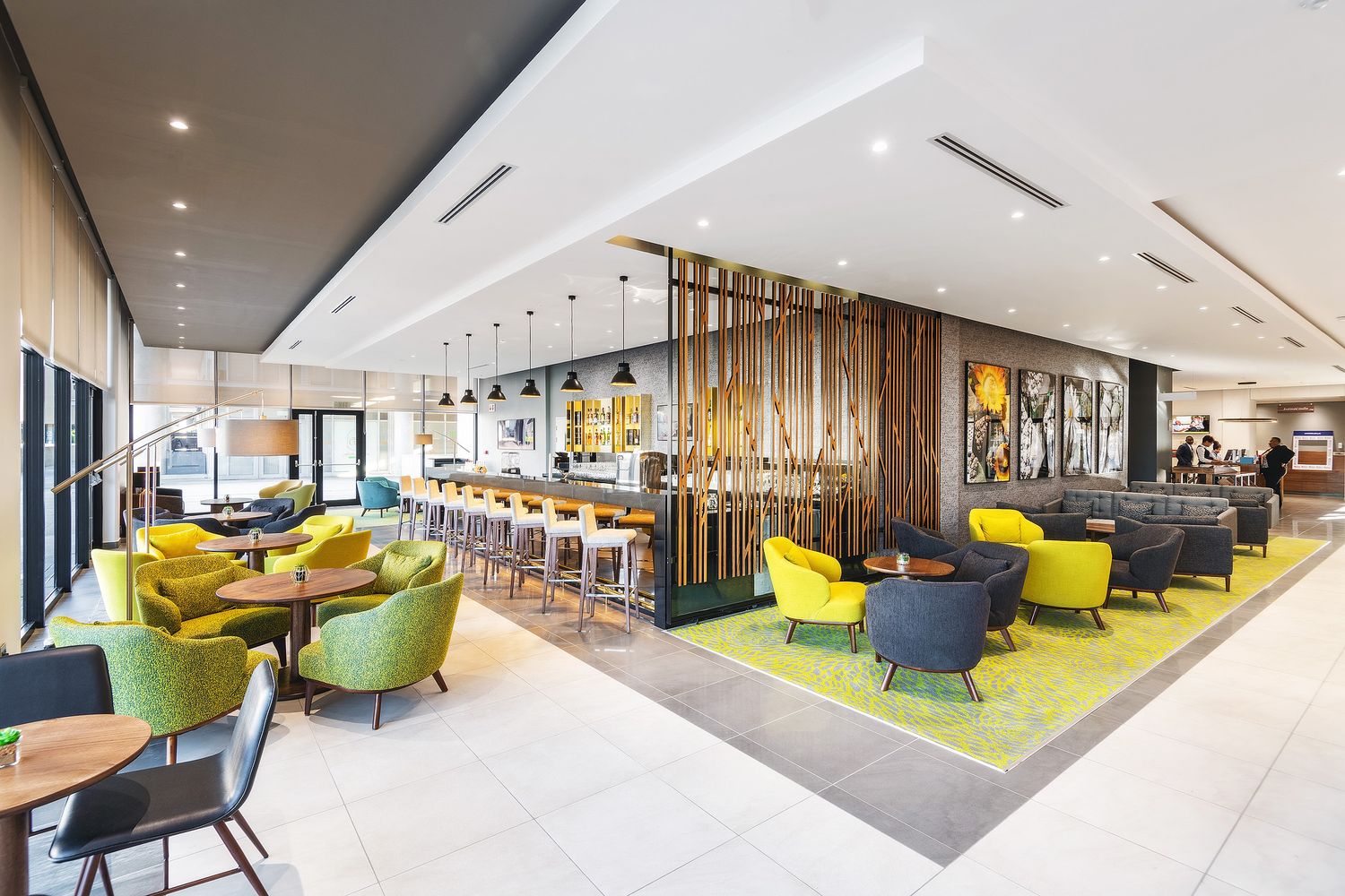 Modern hotel lobby bar with yellow-green chairs, pendant lights, and stylish seating areas on patterned carpet.