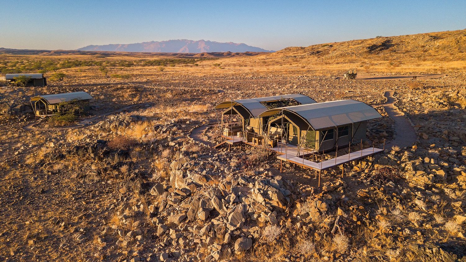 Aerial view of remote desert lodge with green-roofed cabins surrounded by arid landscape and mountain backdrop at sunset.