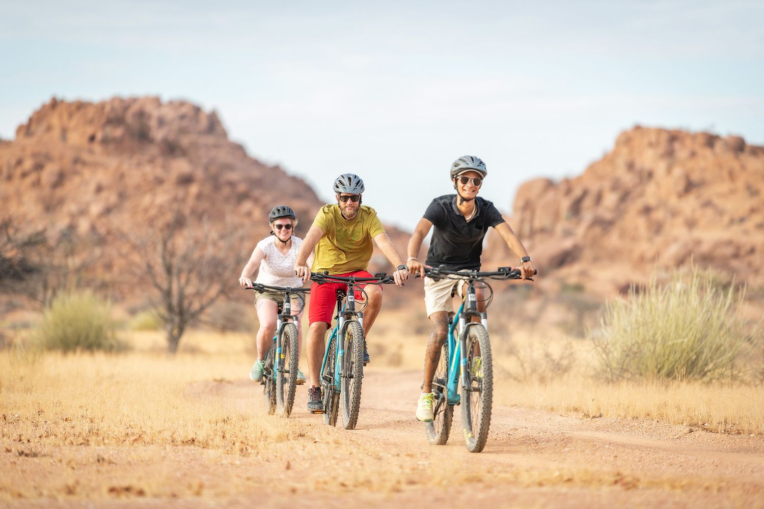 Three cyclists ride mountain bikes on a desert trail with rocky hills in the background under a clear blue sky.