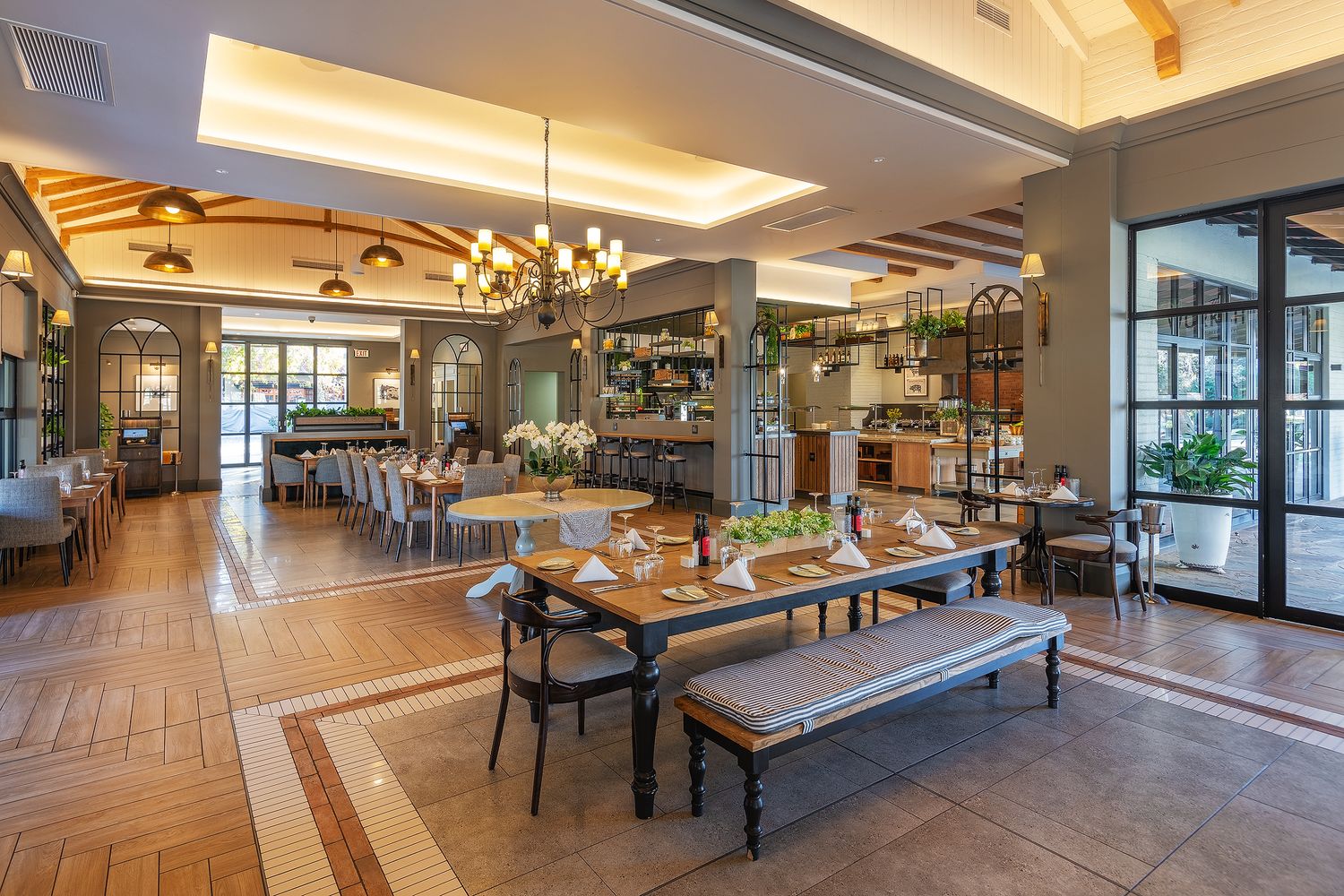 Elegant restaurant interior with wooden tables, bench seating, chandelier lighting, and an open bar area with large windows.