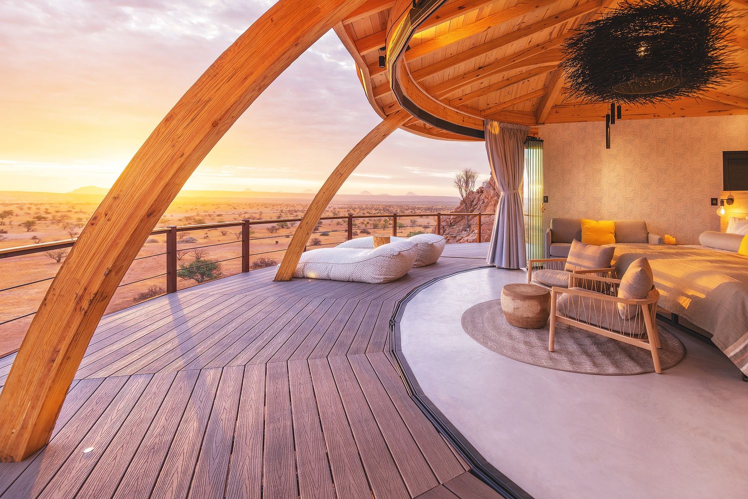 Luxury open-air desert suite with curved wooden arch, outdoor deck bed, and panoramic sunset views.