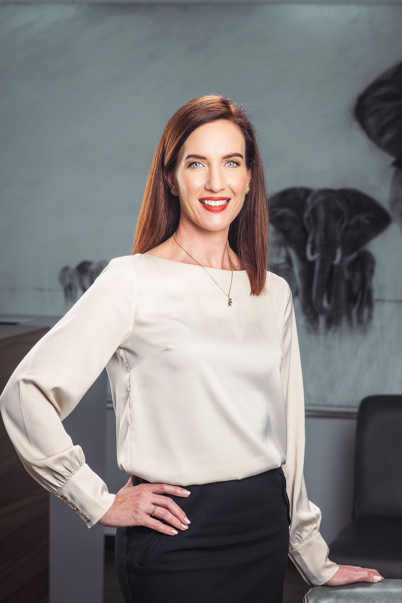Professional woman in white blouse smiling confidently in modern office with elephant artwork background.