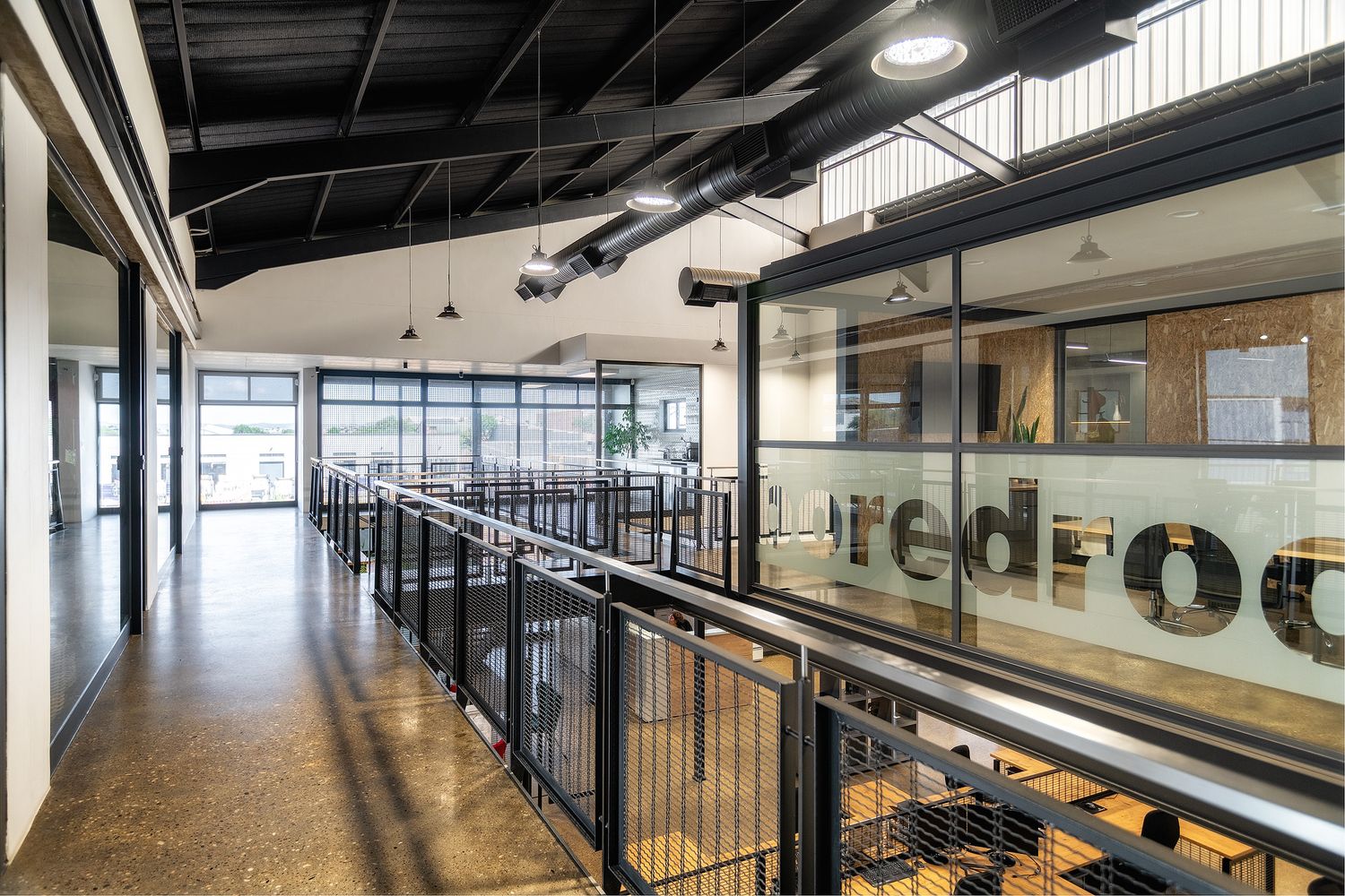Modern office mezzanine with black railings, industrial ceiling, and glass walls featuring red branding logo.