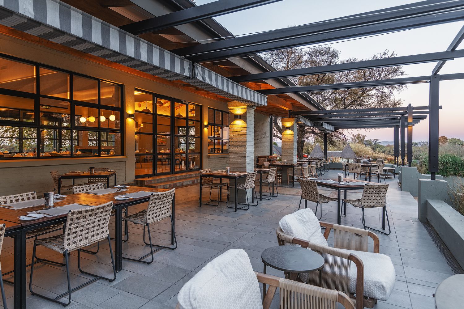 Elegant outdoor restaurant patio with wicker chairs, wooden tables, pergola roof, and warm ambient lighting at dusk.