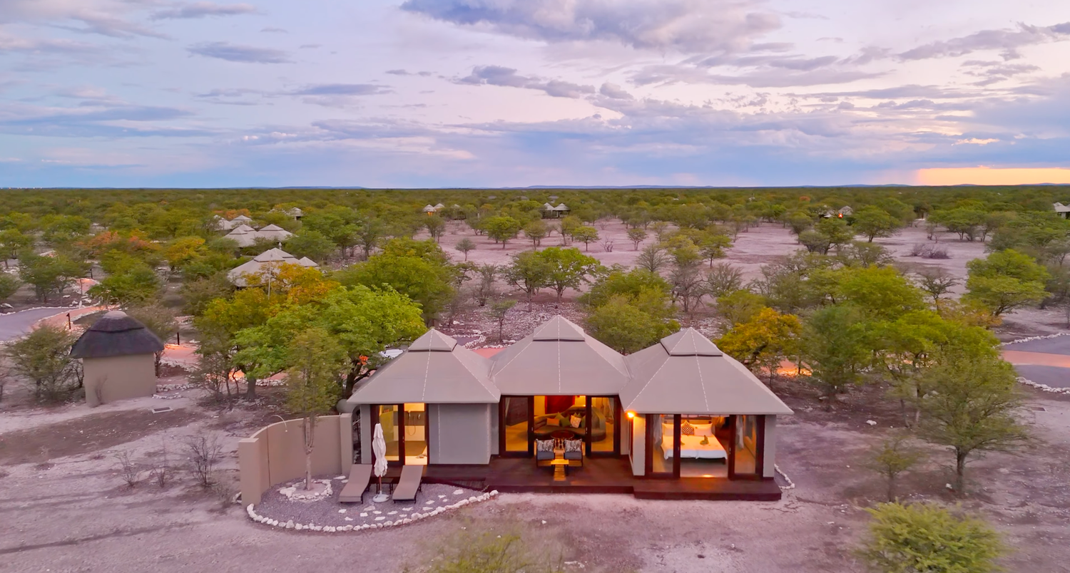 Aerial view of a luxury safari lodge surrounded by African bushveld at dusk, with warm interior lights glowing.