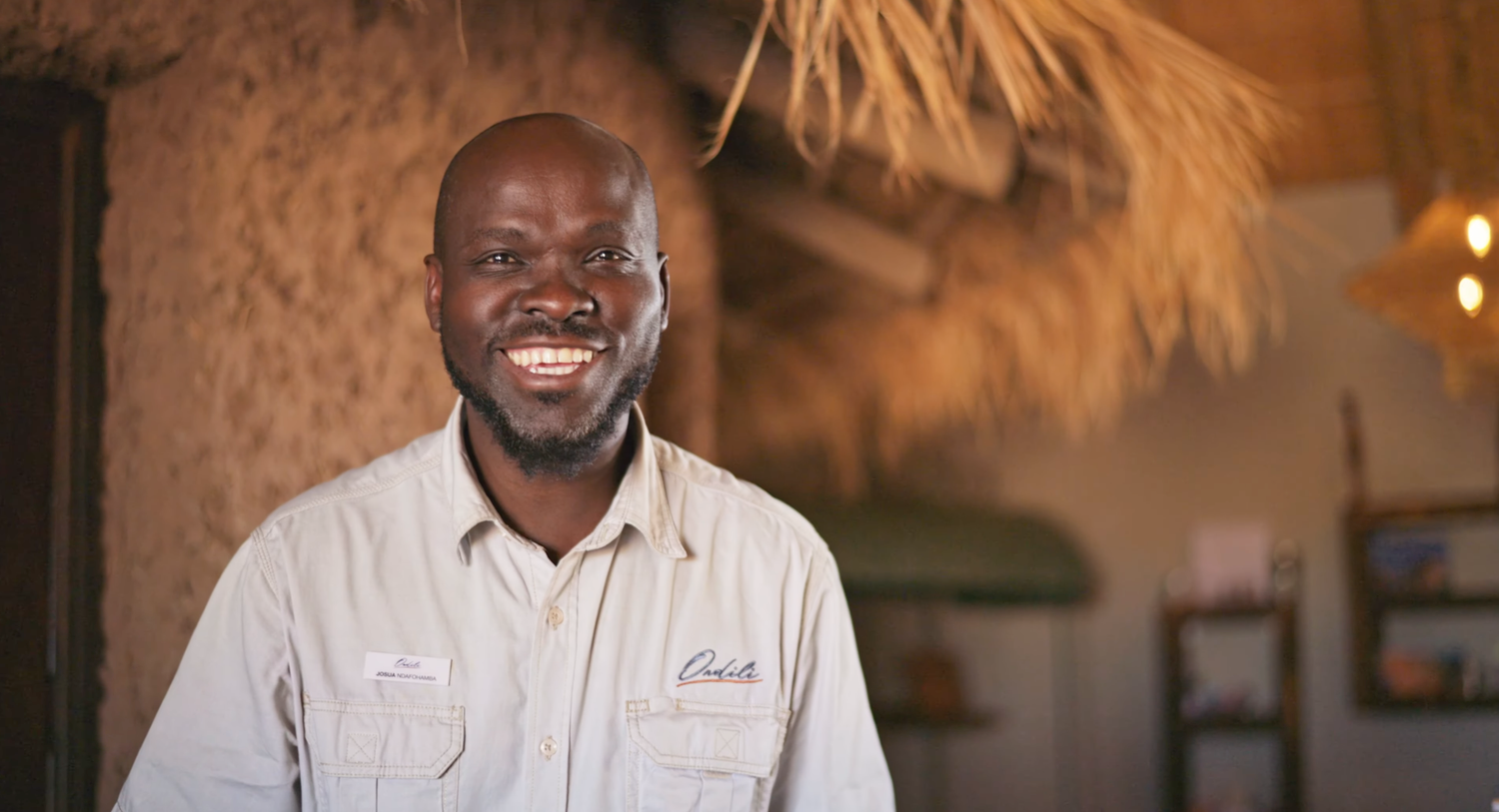 Smiling African man in beige uniform shirt standing in a rustic thatched interior setting.
