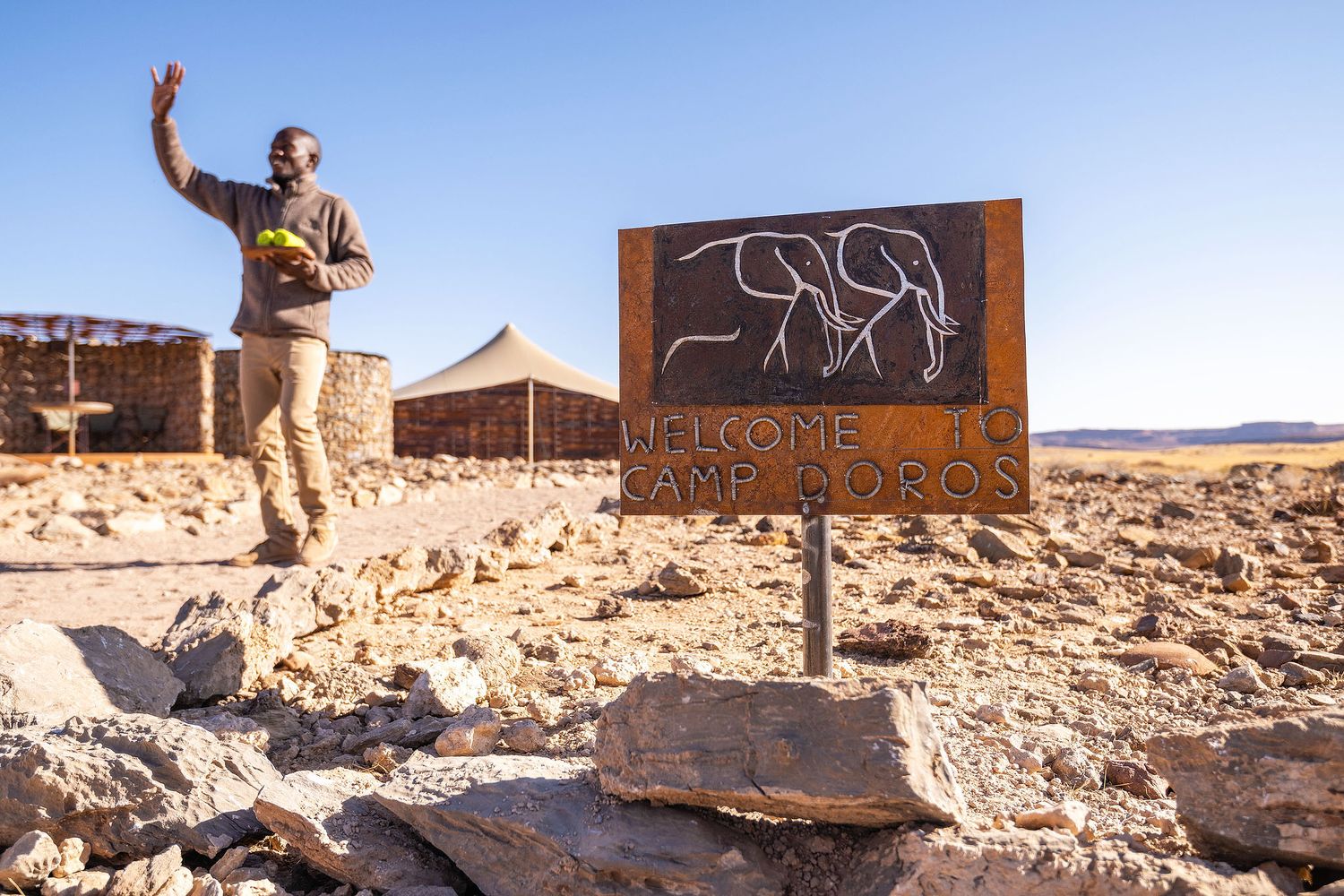 Man waving near a Welcome to Camp Auros sign in a rocky desert landscape under a clear blue sky.