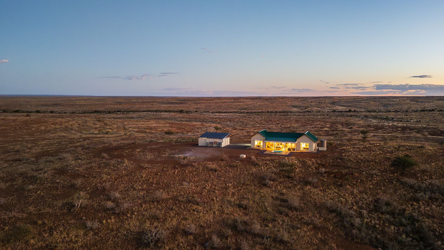Aerial view of an isolated ranch with lit buildings at dusk, surrounded by vast open plains under a twilight sky.