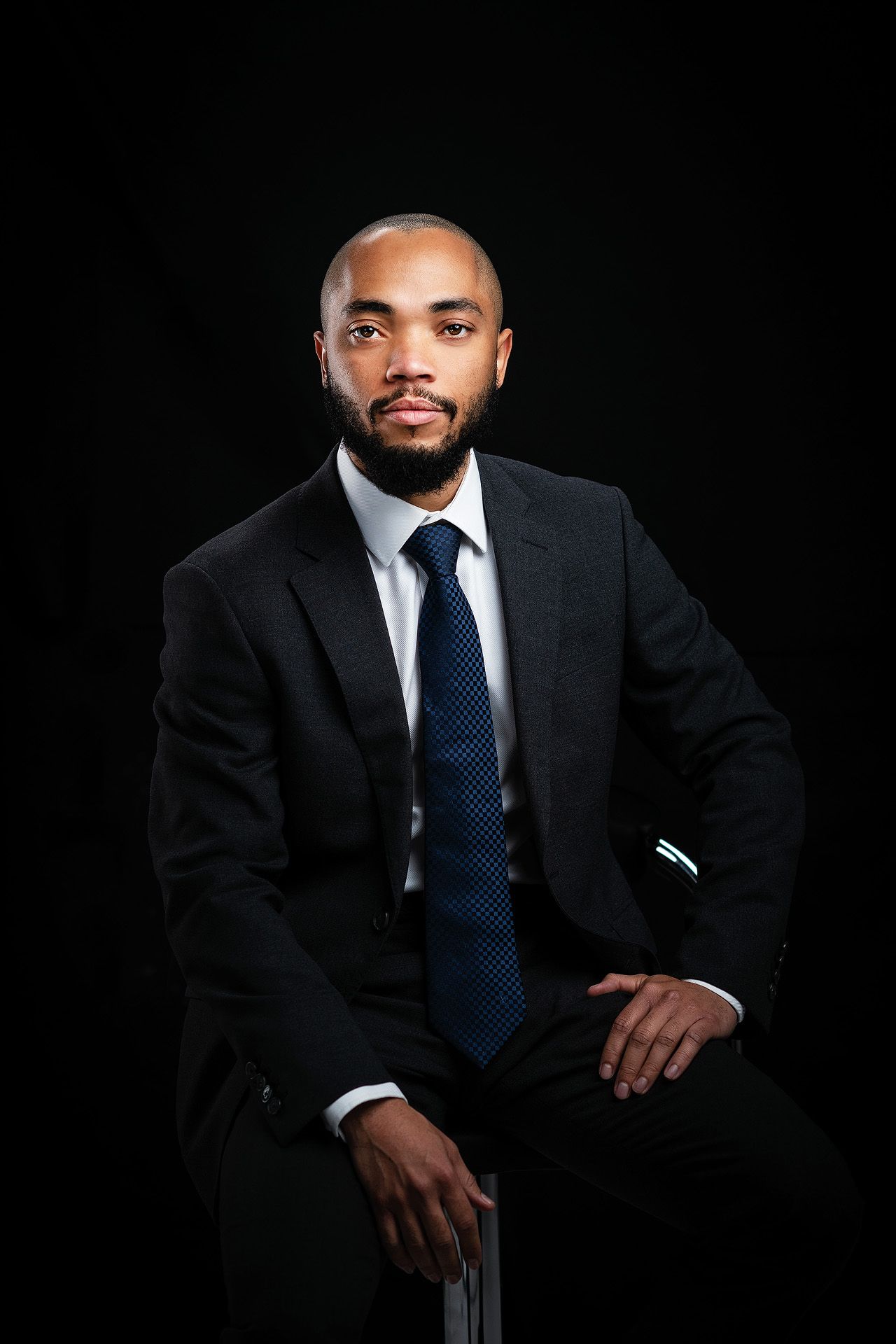 A professional Black man in a dark suit and blue tie sits confidently against a black background.