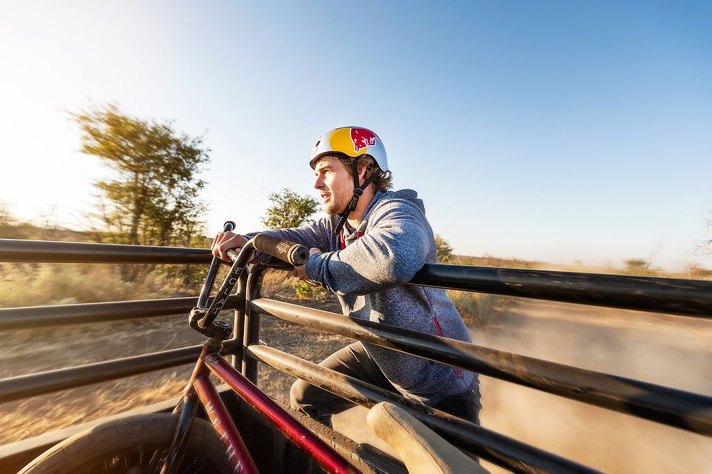 BMX rider wearing Red Bull helmet races along wooden fence track at high speed in sunny outdoor setting.