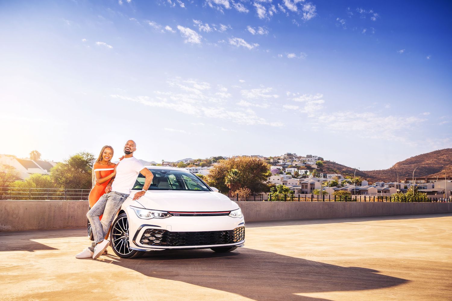 Couple leaning on a white Volkswagen Golf GTI in a sunny parking lot with a scenic hillside village in the background.