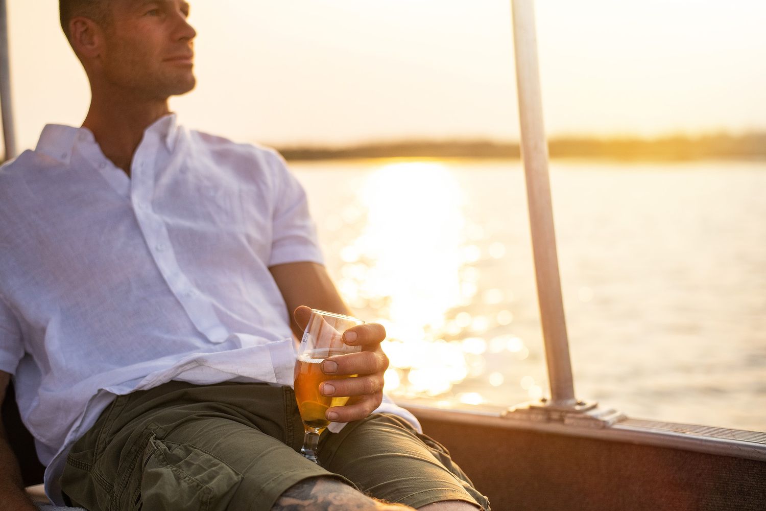 Man relaxing on a boat at sunset holding a glass of beer with golden water reflections in the background.