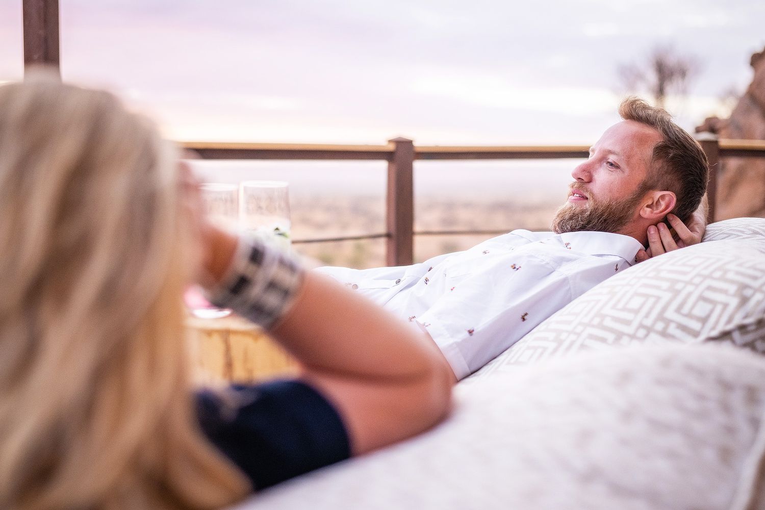 Couple relaxing outdoors on a cozy bed with a scenic view, man lying back thoughtfully while woman holds a drink.