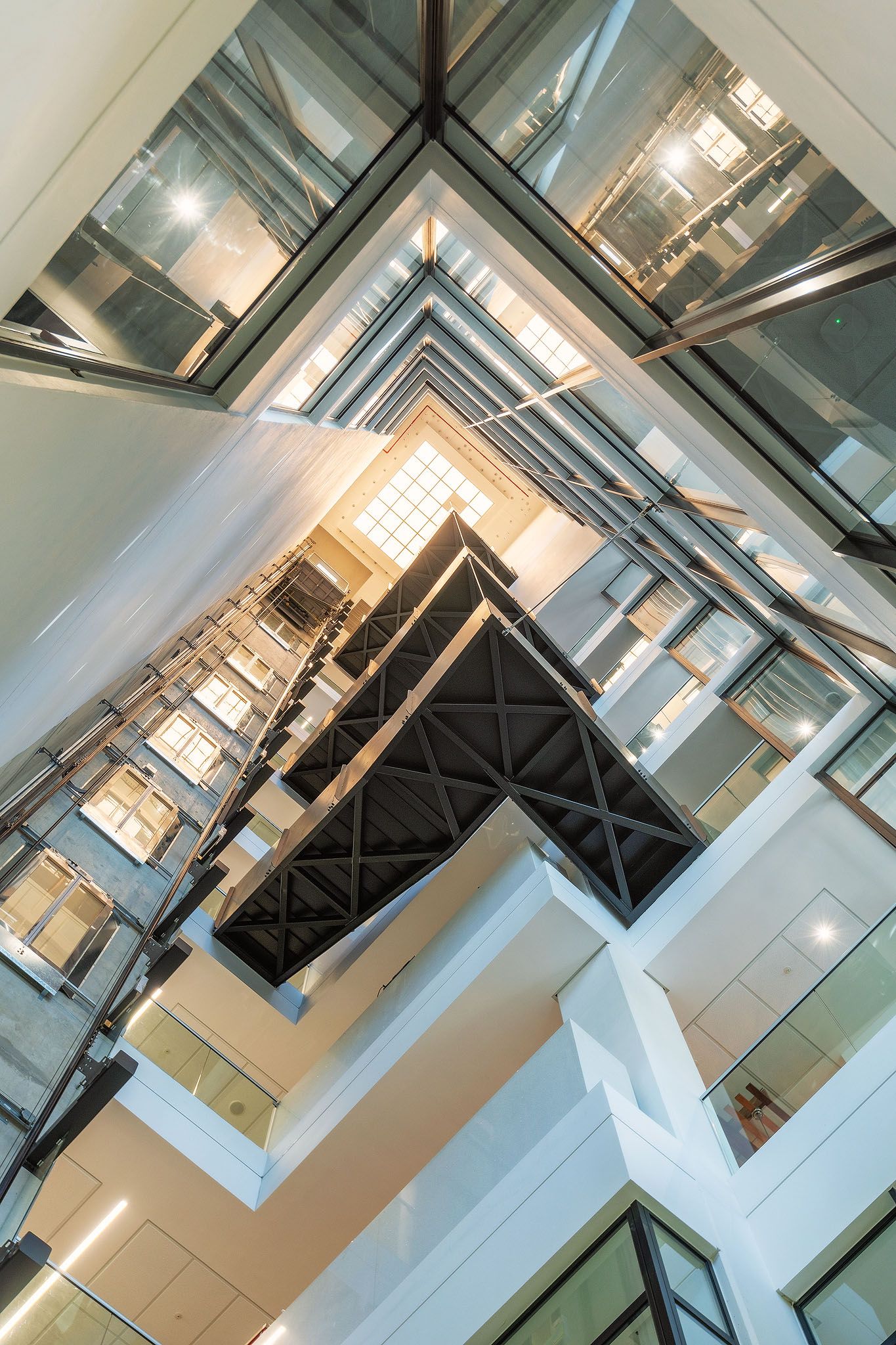 Upward view of geometric glass elevator shaft and staircases in a modern multi-story office building.