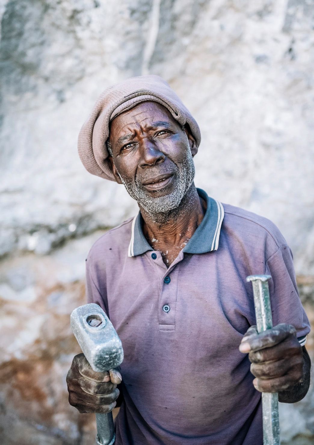 Elderly African man holding two hammers, wearing a pink polo shirt and knit hat, working outdoors near a rock wall.
